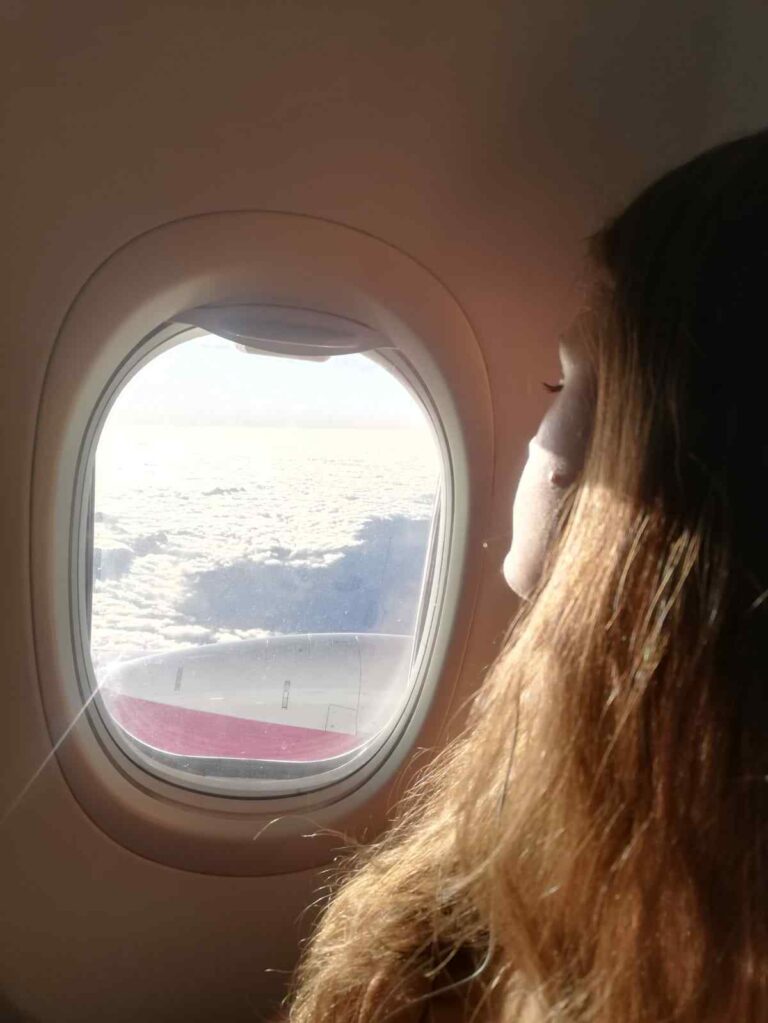 Nikita with long hair sitting at a window seat inside an airplane looking out the window over the clouds.