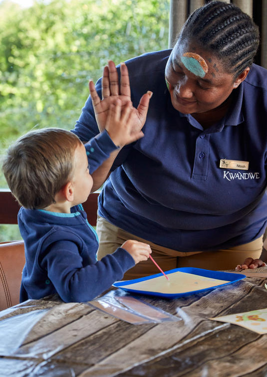 Child-minder at a safari lodge giving a young child a hi-five in the activities room