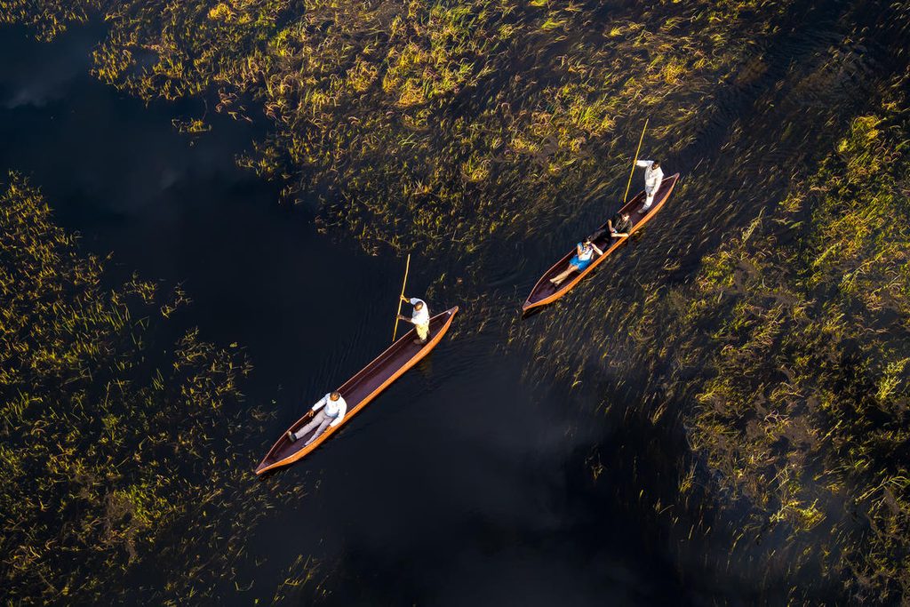 An aerial view of two mokoros in the Okavango Delta - the front mokoro has one person with a poler, the second mokoro has two people in it with a poler.